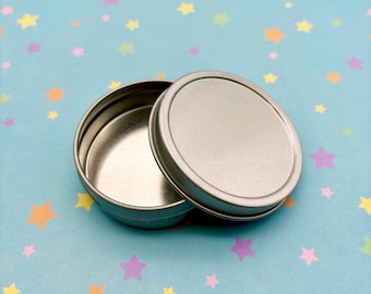 May include: A silver metal tin with a lid. The tin is round and has a smooth surface. The lid is slightly larger than the base of the tin.