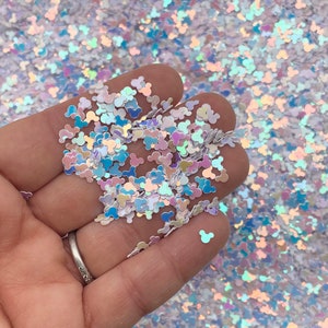 May include: Iridescent glitter in the shape of Mickey Mouse heads. The glitter is a mix of white, blue, and pink, with a shimmering, holographic effect. The glitter is held in a hand, with more glitter in the background.