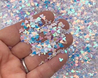 May include: Iridescent glitter in the shape of Mickey Mouse heads. The glitter is a mix of white, blue, and pink, with a shimmering, holographic effect. The glitter is held in a hand, with more glitter in the background.