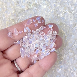 May include: A close-up of crushed opal gemstones, with a milky white base and flashes of colour. The small, irregular pieces are held in a hand, with more scattered in the background. The gemstones have an iridescent quality.