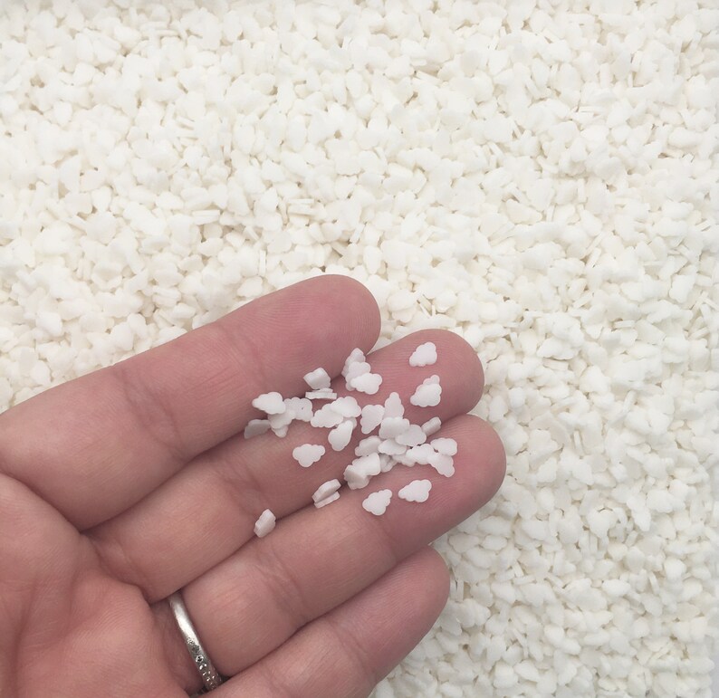 May include: A hand holding a small pile of white cloud-shaped sprinkles. The background is filled with similar white, irregular-shaped sprinkles. A silver ring is visible on the person's finger.