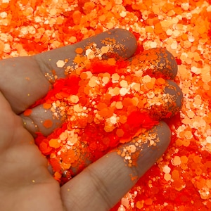 Puede incluir: Primer plano de una mano sosteniendo un montón de purpurina naranja brillante. La purpurina está compuesta por pequeñas piezas hexagonales.