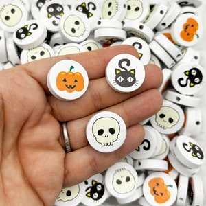 Può includere: Una collezione di perline rotonde in legno bianco con disegni a tema Halloween. I disegni includono un gatto nero, una zucca e un teschio.
