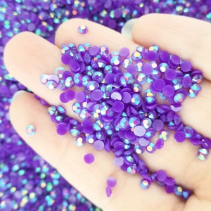 May include: Close-up of a hand holding a pile of purple, iridescent rhinestones. The rhinestones are round and flat-backed, with a glossy finish. The background is filled with more rhinestones, creating a sparkling effect. Ideal for crafts and embellishments.