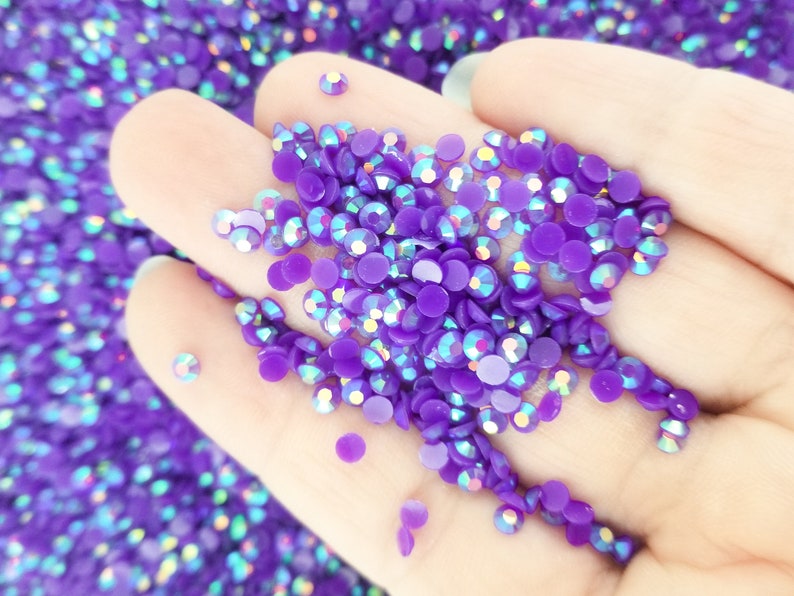 May include: Close-up of a hand holding a pile of purple, iridescent rhinestones. The rhinestones are round and flat-backed, with a glossy finish. The background is filled with more rhinestones, creating a sparkling effect. Ideal for crafts and embellishments.