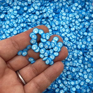 May include: A close-up of a hand holding a pile of blue and white resin bubble charms. The charms are small and round, with a white outline and a blue center. The charms are perfect for adding a touch of whimsy to any craft project.