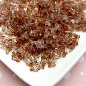 以下が含まれることがあります： 茶色と黒の羽根を持つ、小さな透明なプラスチック製の蝶のチャームが、白い皿の上に散らばっています。
