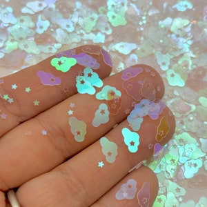 May include: A close-up of iridescent holographic cloud and star shaped glitter. The glitter is scattered on a hand and a table.