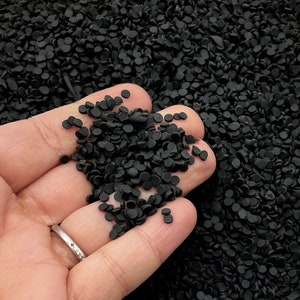 Könnte beinhalten: Eine Hand hält eine Ansammlung kleiner, schwarzer, kreisförmiger Objekte. Die Objekte sind in der Hand verteilt und scheinen aus einem matten Material zu bestehen. Der Hintergrund ist mit weiteren schwarzen Objekten gefüllt, was eine dichte Textur erzeugt.