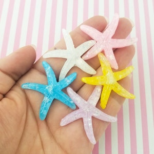 May include: Five glittery starfish in various colors, including pink, white, blue, and yellow. The starfish are arranged in a hand, with the tips of the starfish pointing outwards.