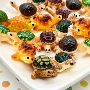 May include: A collection of miniature turtle figurines in various colors, including green, brown, and yellow, are arranged on a white rectangular plate. The turtles have detailed shells and visible eyes, creating a playful and decorative display.
