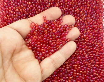 May include: A hand holding a handful of small, red, iridescent beads. The beads are scattered across a surface.