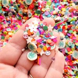 May include: A hand holding a variety of colorful sprinkles, including stars, flowers, smiley faces, and leaves. The sprinkles are made of plastic and are perfect for decorating cakes, cupcakes, and other desserts.