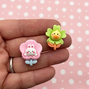 May include: Two colourful resin flower charms. One charm is pink with a white bunny face and the other is green with an orange duck face.
