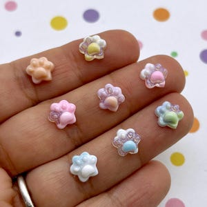 Könnte beinhalten: Sortiment an bunten Harz-Pfotenabdruck-Anhängern mit Glitzerakzenten. Die Anhänger sind in verschiedenen Pastellfarben, darunter Rosa, Blau, Gelb und Grün.