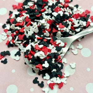 May include: A close-up of red, black, and white sprinkles shaped like Mickey Mouse heads. The sprinkles are scattered on a pink surface with white polka dots.