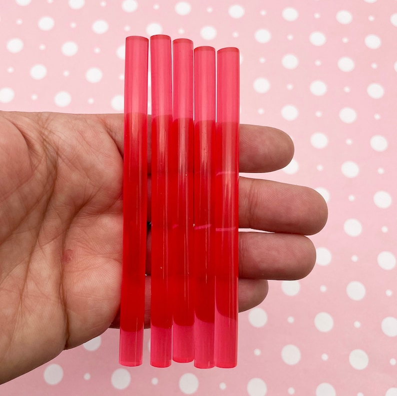 May include: Five translucent red glue sticks, approximately 4 inches long, are held in a hand. The glue sticks are arranged in a row, with the ends of the sticks aligned.