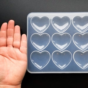 May include: A clear silicone mold with nine heart-shaped cavities. The mold is on a black surface.