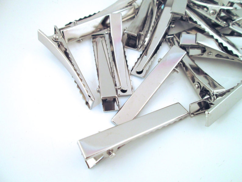 Puede incluir: Un mont&oacute;n de pinzas para el pelo de cocodrilo de metal plateado. Las pinzas son todas del mismo tama&ntilde;o y forma, y est&aacute;n dispuestas en un patr&oacute;n aleatorio.