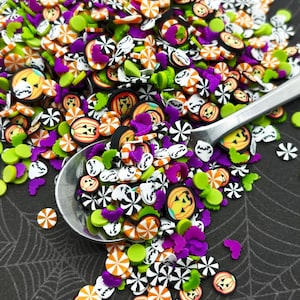 May include: A variety of Halloween-themed sprinkles in a silver spoon. The sprinkles are in the shape of pumpkins, candy canes, bats, and ghosts. The colors are orange, green, purple, black, and white.