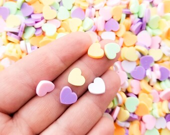 May include: A hand holding a variety of small heart-shaped candies in pastel colors, including pink, purple, yellow, and white. The candies are scattered on a surface.