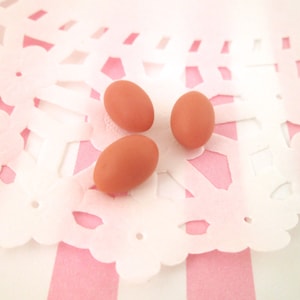 Può includere: Tre piccole uova ovali, marrone-arancio, sono disposte su un centrino bianco a motivi floreali. Il centrino poggia su uno sfondo a righe rosa e bianche, creando un'immagine dai toni pastello tenui. Le uova appaiono lisce e piene.