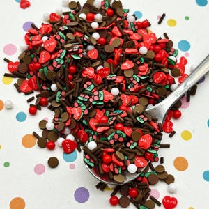 Op de afbeelding: Een verzameling decoratieve strooisels met bruine staafjes, kleine witte bolletjes, rode hartvormige stukjes met de tekst LOVE en groen-rode kersvormige figuurtjes, allemaal opgeschept met een zilverkleurige metalen lepel.