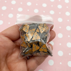 May include: A clear plastic bag filled with small brown and grey resin poop emoji charms. The charms have a smiley face with two black eyes.