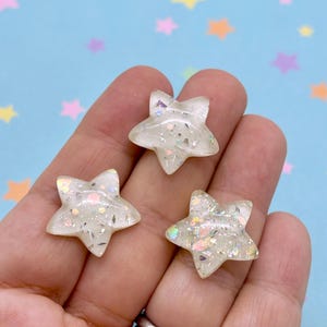 May include: Three translucent star-shaped embellishments with iridescent glitter flecks. The stars are held in a hand against a light blue background decorated with colorful star shapes. These decorative stars are ideal for crafts and DIY projects.