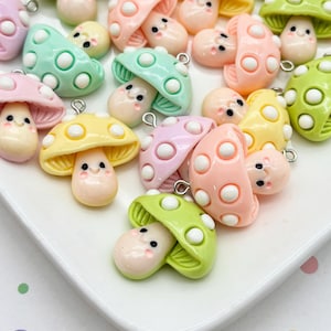 May include: A collection of small, glossy mushroom charms in pastel shades of pink, yellow, green, and lavender. Each mushroom features a smooth, rounded cap with white polka dots, a smiling face with black eyes, and a small metal jump ring at the top.