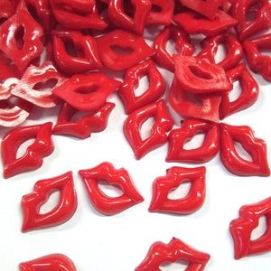 Puede incluir: Un conjunto de cuentas brillantes en forma de labios, de color rojo vivo, dispersas sobre una superficie blanca. Las cuentas tienen un acabado liso y reflectante, ideales para manualidades o joyería.