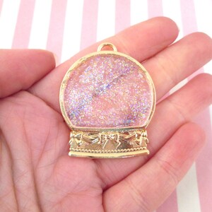 May include: Gold-tone metal snow globe charm with a clear dome filled with pink glitter. The charm has a decorative base with a bow design.