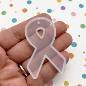 May include: A clear, ribbon-shaped mold with a small hole at the top. The mold is held in a hand against a background of colorful polka dots. The ribbon design is a symbol of awareness and support.