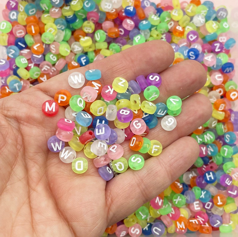 May include: A close-up of a hand holding a variety of colourful alphabet beads. The beads are translucent and have white letters on them. The beads are in a variety of colours, including pink, blue, green, yellow, and orange.