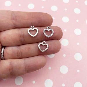May include: Three silver heart-shaped charms with a loop at the top. The charms are on a pink background with white polka dots.