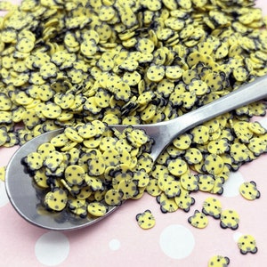 Op de afbeelding: Een lepel gevuld met gele en zwarte kattengezichtjes sprinkles. De sprinkles zijn verspreid over een roze oppervlak met witte stippen.