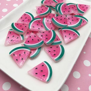 May include: A pile of pink and green watermelon slice shaped resin charms with glitter. The charms are on a white plate with a pink polka dot background.