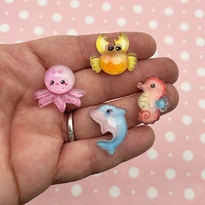 May include: Four clear resin sea creature charms. A pink octopus, a yellow crab, a blue dolphin, and an orange seahorse.