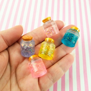 May include: A collection of miniature, clear jars filled with colorful glitter and small objects. Each jar has a wooden lid and a yellow band. The jars are held in a hand against a pink and white striped background.