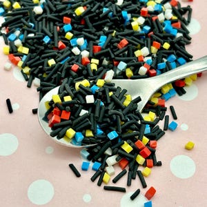Op de afbeelding: Zwarte, rode, blauwe en gele sprinkles in verschillende vormen, waaronder lange stokjes en kleine vierkantjes. De sprinkles zijn verspreid over een roze oppervlak met witte stippen.