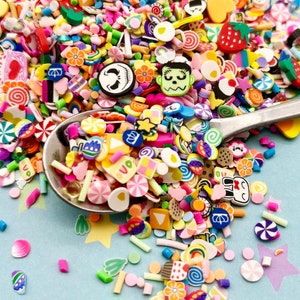 Pode incluir: Uma variedade de confeitos de argila polimérica coloridos, incluindo doces, flores e personagens de desenhos animados. Os confeitos estão em uma colher e espalhados em um fundo azul.