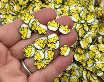 Pode incluir: Um close-up de uma mão segurando uma variedade de enfeites de resina em forma de pintinhos amarelos e brancos. Os pintinhos estão em uma casca de ovo rachada com contornos pretos e bicos laranja.