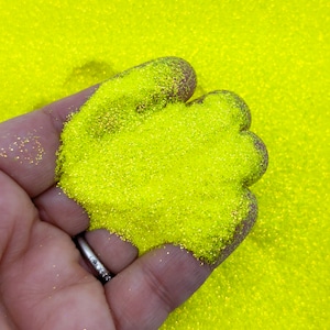 May include: A close-up of a hand holding a pile of bright yellow glitter. The glitter is fine and sparkly, and it is perfect for crafting or decorating.
