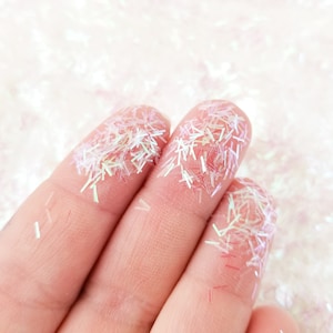 以下が含まれることがあります： 2 本の指に虹色の白いキラキラした粉がまぶされているクローズアップ。キラキラは小さく、薄い小さな破片に似ています。