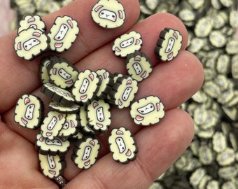 May include: A hand holding a variety of small, light brown and cream-colored resin sheep charms. The charms have black outlines and white faces with pink cheeks.