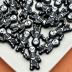 Può includere: Una collezione di piccoli abbellimenti a forma di scheletro nero con dettagli bianchi, disposti su un piatto bianco. Gli scheletri hanno un design a cartone animato, con gabbie toraciche e teschi visibili. Lo sfondo è una superficie arancione chiaro. Questi sono probabilmente materiali per l'artigianato.