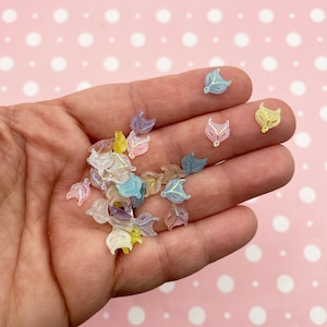 Puede incluir: Un puñado de pequeños adornos translúcidos en forma de hoja en varios colores pastel, incluyendo azul, morado y amarillo. Cada hoja tiene un delicado diseño de vetas brillantes. El fondo es una superficie rosa con lunares blancos.