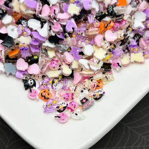 Puede incluir: Una colección de adornos de resina con temática de Halloween en varias formas y colores. El surtido incluye fantasmas, calabazas, gatos, murciélagos y sombreros de bruja en tonos rosa, morado, negro, blanco y naranja. Los adornos están esparcidos sobre una superficie blanca.