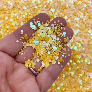 May include: A close-up of a hand holding a handful of yellow iridescent glitter. The glitter is in various sizes and shapes, and it is scattered on a surface.