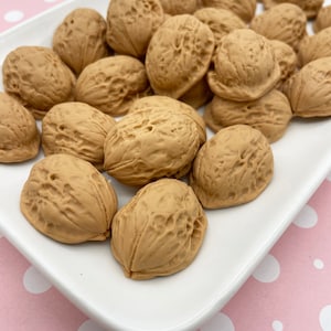 Könnte beinhalten: Ein weißer Keramikteller gefüllt mit einem großen Haufen brauner Walnüsse. Der Teller befindet sich auf einem rosa Hintergrund mit weißen Punkten.